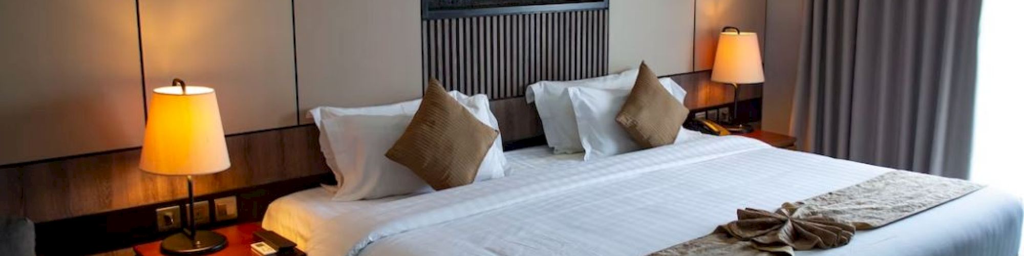 A modern hotel room with a large bed, two side tables with lamps, decorative pillows, and a wall art piece. Linen is neatly arranged on the bed.