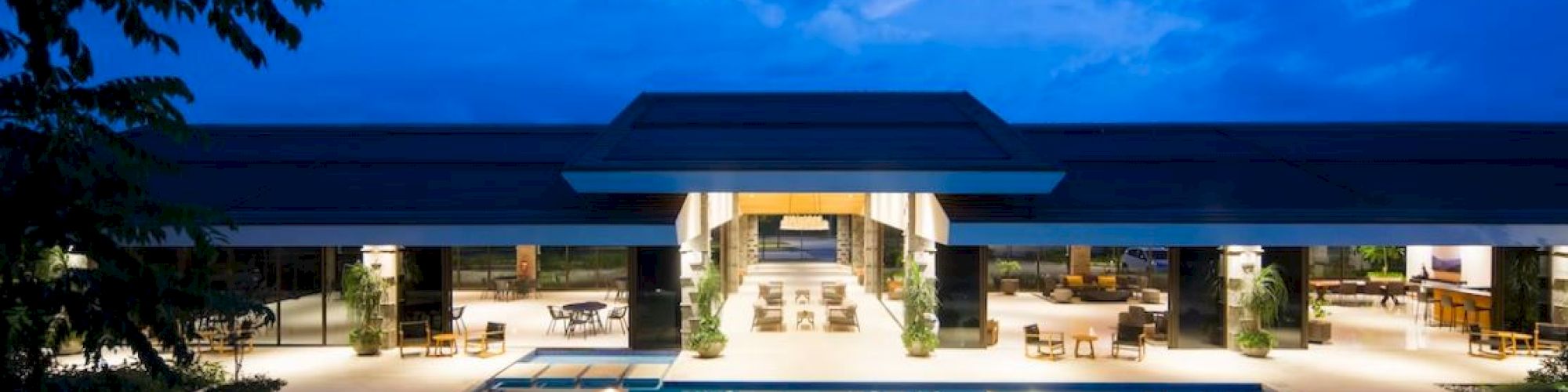 A luxurious modern building with a large, illuminated outdoor pool in front, surrounded by trees under a deep blue evening sky.