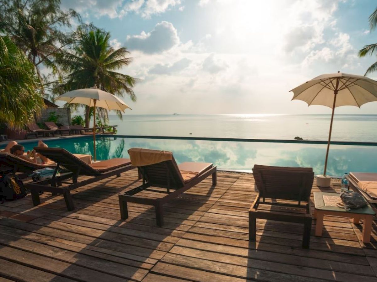 A sunlit poolside with lounge chairs, umbrellas, and palm trees, overlooking a serene seascape under a partly cloudy sky.