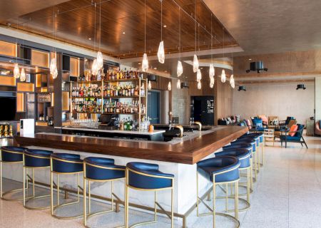 A modern bar with blue padded stools, hanging lights, and a well-stocked drink shelf; cozy seating area in the background completes the space.