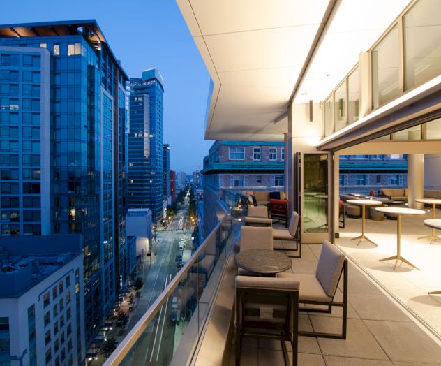 A modern balcony with seating overlooks a cityscape with tall buildings and a street illuminated at dusk.