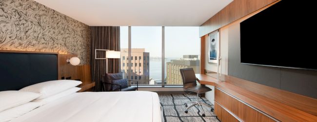 Modern hotel room with large bed, wall-mounted TV, desk, and a cityscape view through large windows.