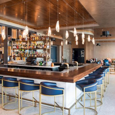 A modern bar with a wooden counter, blue chairs, and hanging lights. Shelves filled with bottles are in the background. Cozy seating area.