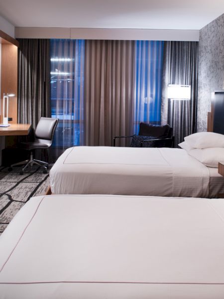 A modern hotel room with two beds, a TV, desk, chair, and lamp. Neutral colors and abstract-patterned carpet complete the look.