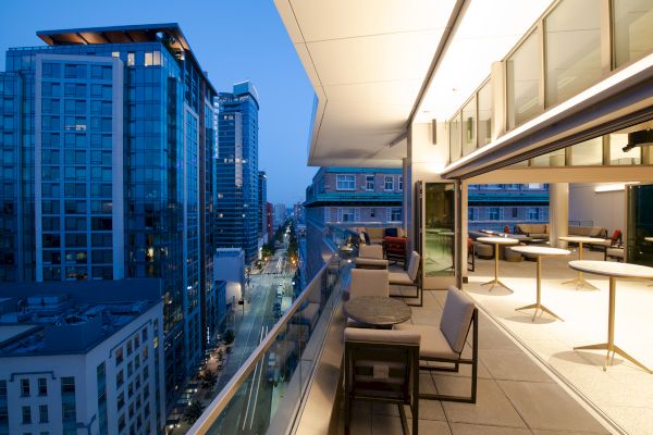 A modern balcony with seating overlooks a cityscape of tall buildings at dusk, with streetlights illuminating the scene.