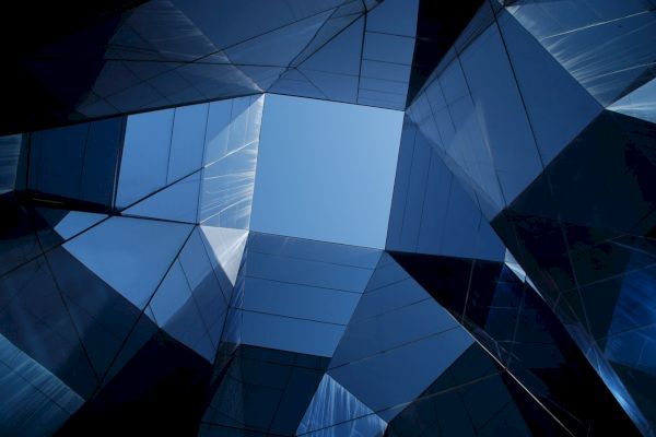 Geometric architectural structures with mirrored surfaces reflect a bright blue sky in the center, creating an abstract visual effect.
