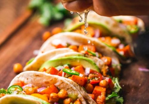 A hand squeezes lime over a row of chickpea and vegetable tacos, garnished with avocado and cilantro on a wooden board.