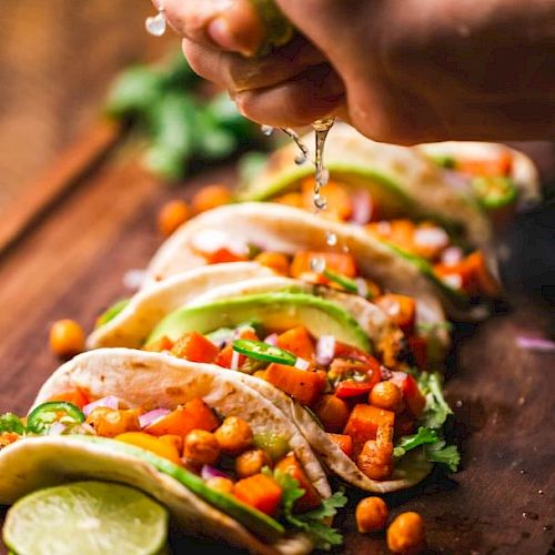 A hand squeezes lime over a row of chickpea and vegetable tacos, garnished with avocado and cilantro on a wooden board.