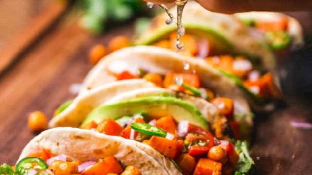 A hand squeezes lime over a row of chickpea and vegetable tacos, garnished with avocado and cilantro on a wooden board.