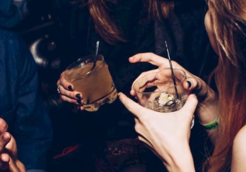 People are enjoying drinks at a social gathering, laughing and gesturing with their hands.