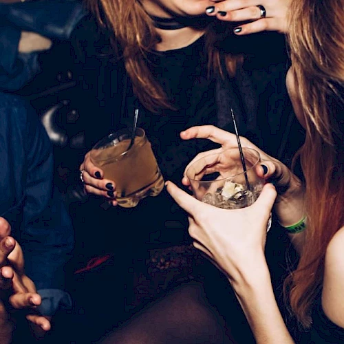People are enjoying drinks at a social gathering, laughing and gesturing with their hands.