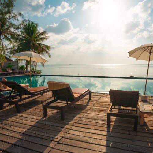 A serene poolside scene with lounge chairs, umbrellas, and palm trees overlooking a calm sea, under a bright sky.