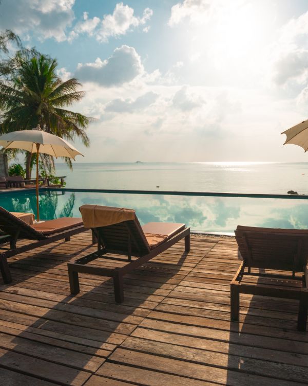 A serene poolside scene with lounge chairs, umbrellas, and palm trees overlooking a calm sea, under a bright sky.