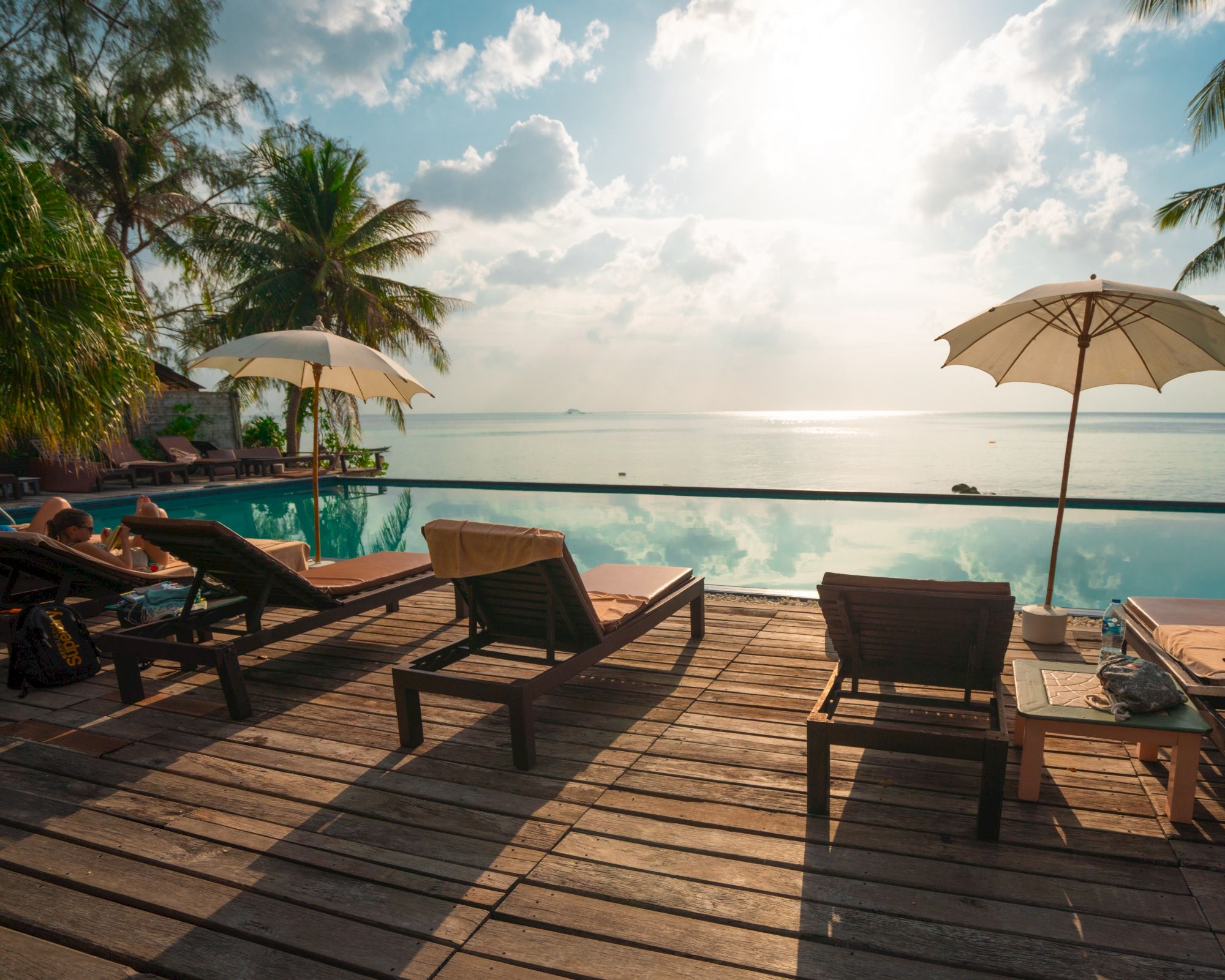 A serene beachside view with wooden lounge chairs, parasols, and palm trees facing a calm ocean under a partly cloudy sky.