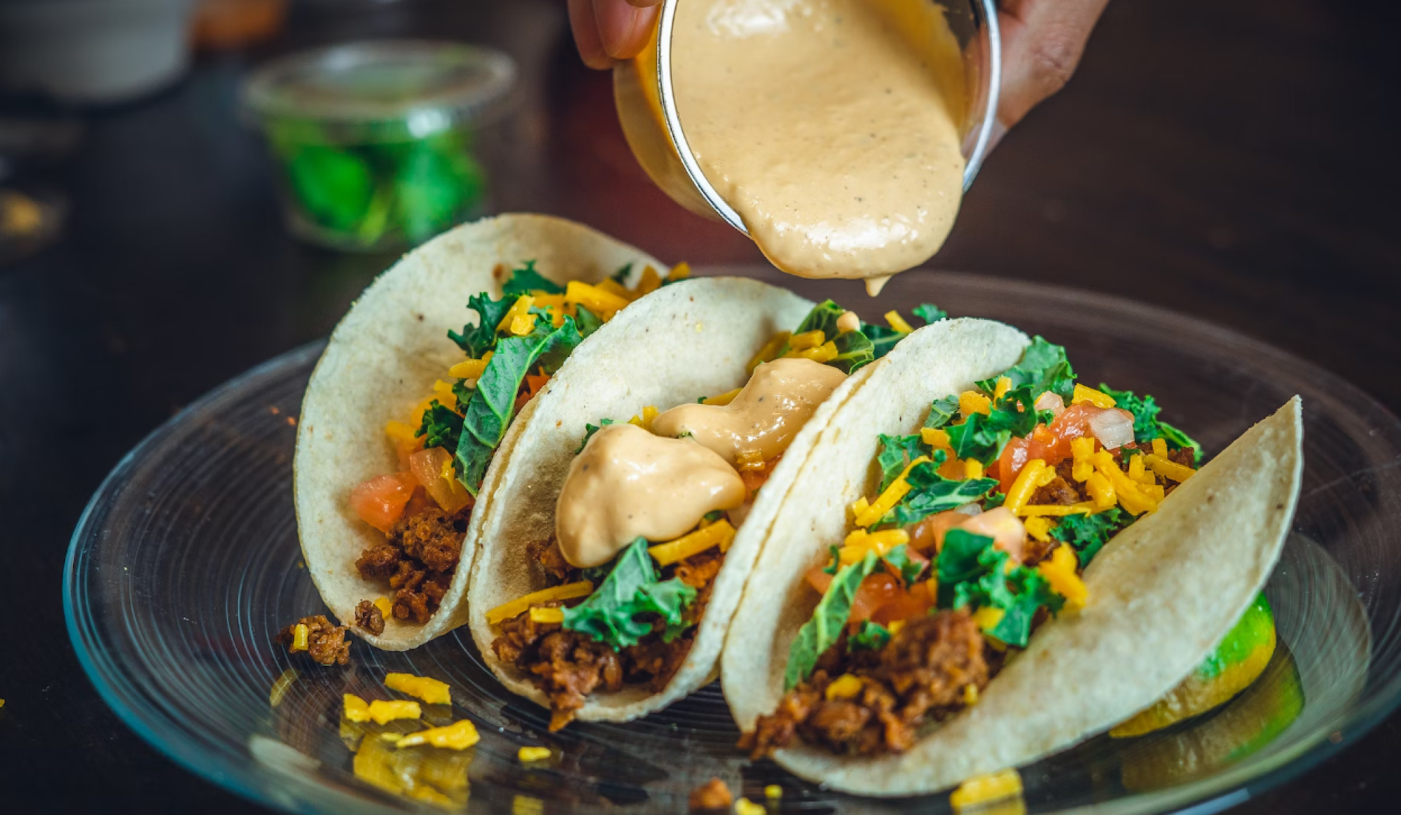 Three tacos with meat, cheese, lettuce, and tomatoes being topped with a creamy sauce on a plate.