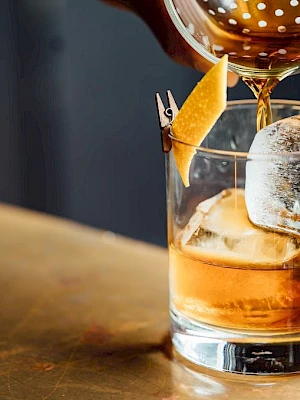 A drink being poured into a glass with a large ice cube and a twist of citrus on the side, set on a countertop.