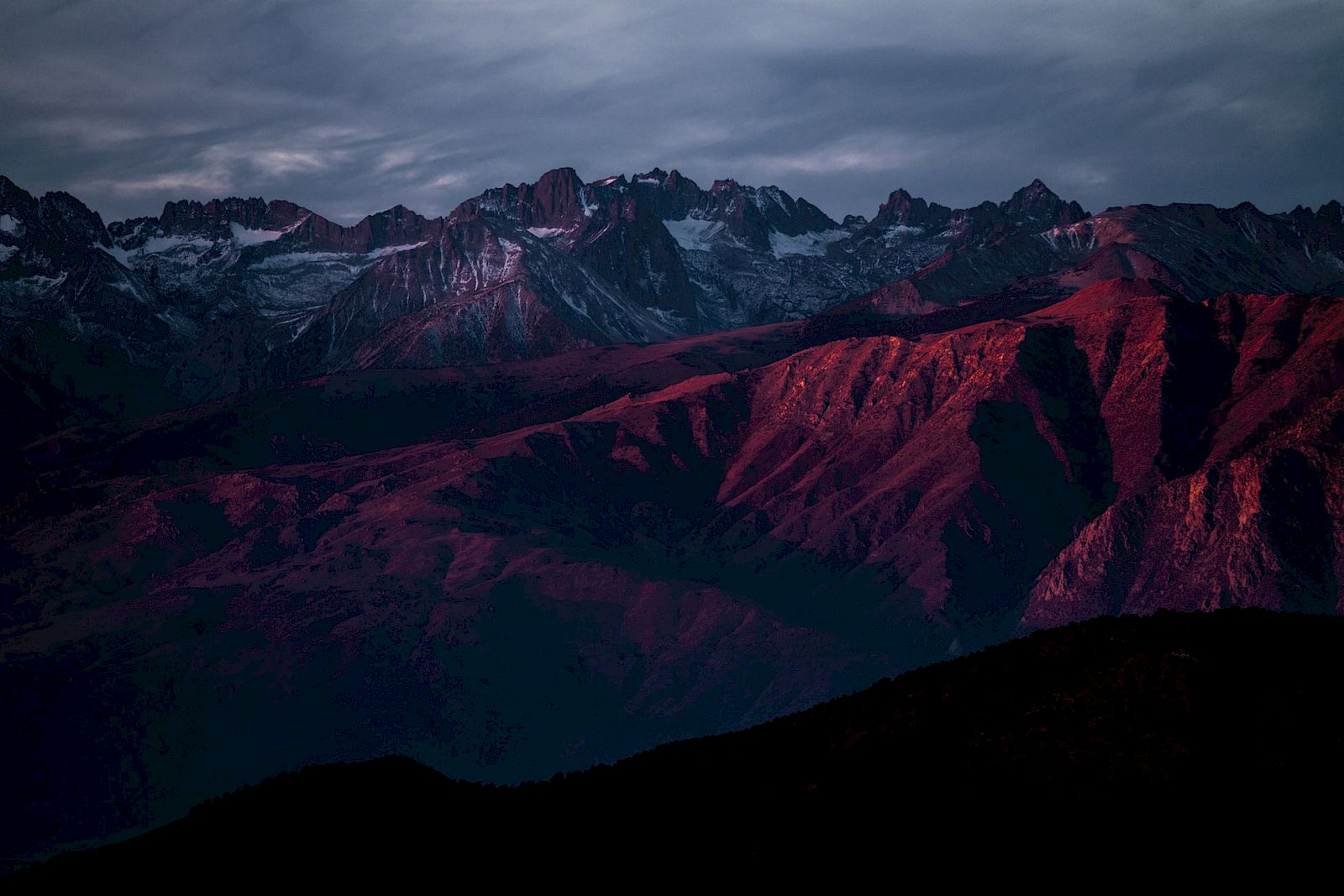 A mountain range is bathed in a warm, red sunset glow under a cloudy sky, creating a dramatic and serene landscape.