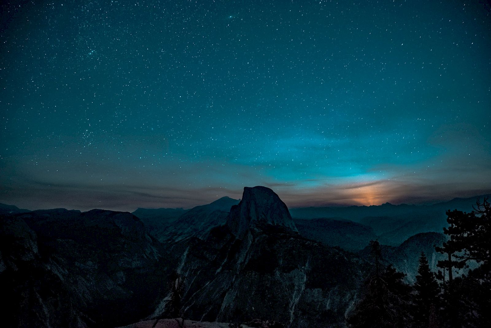 A starlit night sky over a mountain range, with silhouettes of trees in the foreground and a moon rising behind the peaks.