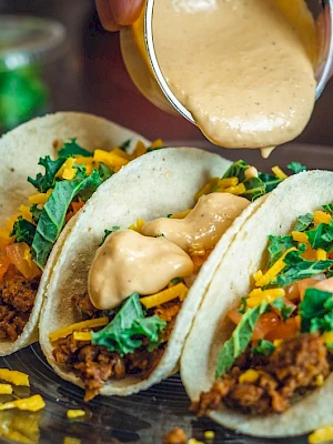 Three tacos with ground meat, lettuce, tomatoes, cheese, and sauce being poured on top, on a plate.