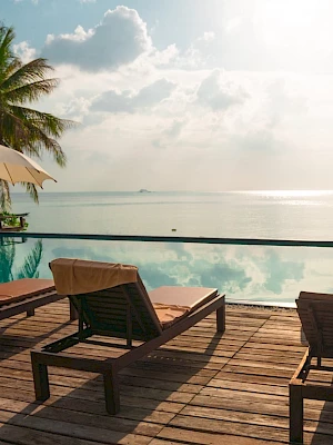 Poolside scene with lounge chairs, umbrellas, and palm trees overlooking a calm sea under a partly cloudy sky.