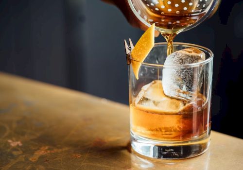 A drink being poured into a glass with a large ice cube and a lemon twist garnish on a wooden surface.