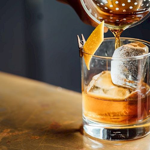A drink being poured into a glass with a large ice cube and a lemon twist garnish on a wooden surface.
