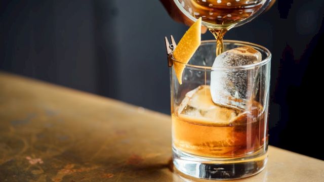 A drink being poured into a glass with a large ice cube and a lemon twist garnish on a wooden surface.