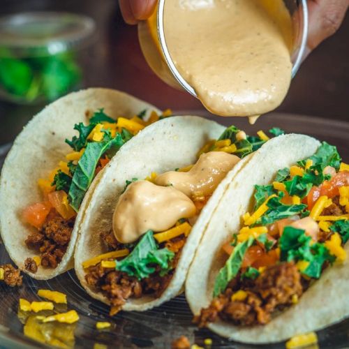Three tacos filled with ground meat, shredded cheese, lettuce, and tomato, topped with a creamy sauce being poured on them.