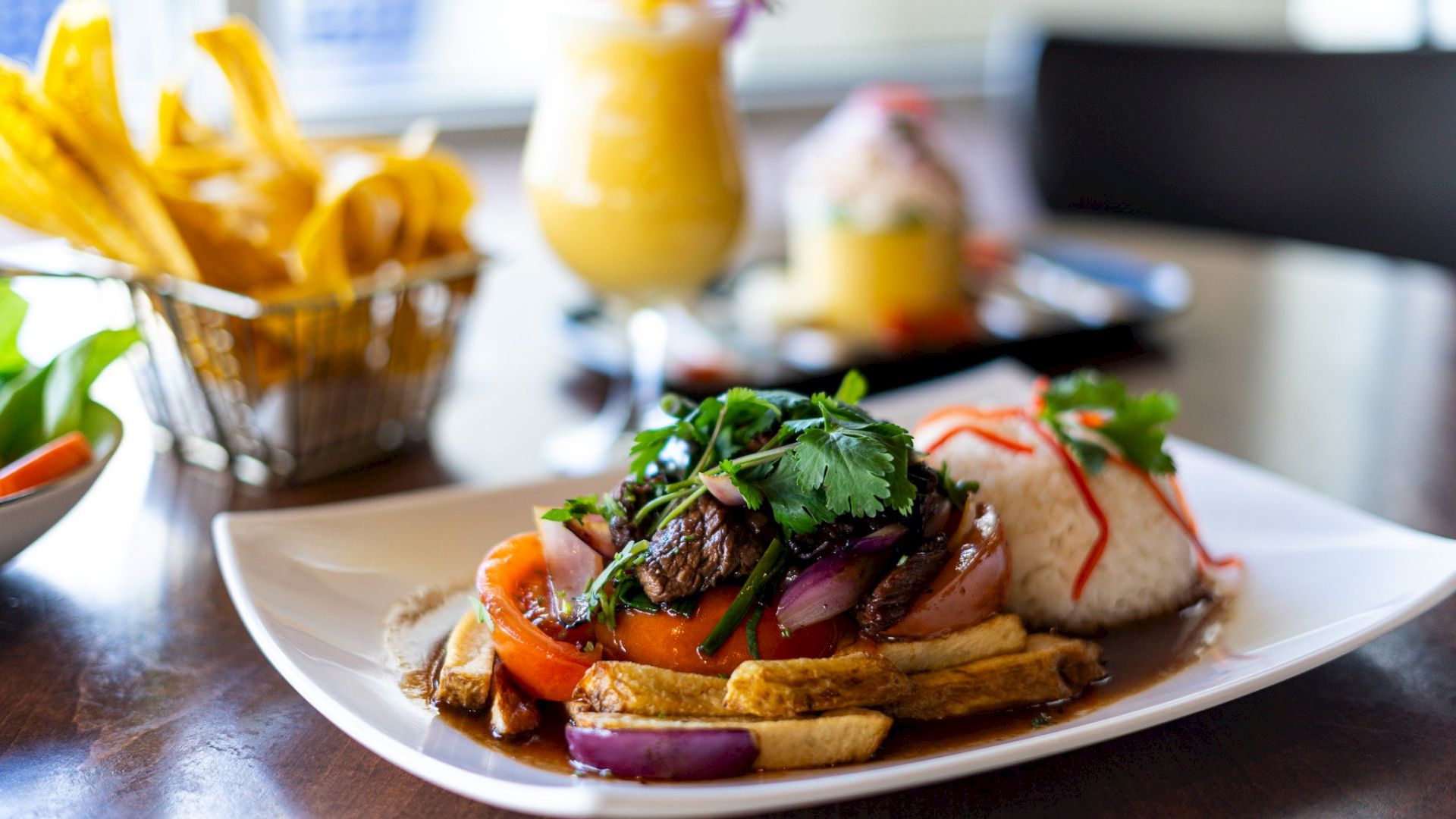 A plate of Lomo Saltado with rice, garnished with cilantro, alongside a basket of fries and two drinks in the background.
