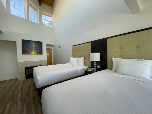 This image shows a hotel room with two beds, a lamp, a painting on the wall, and a high ceiling with large windows and wood flooring.
