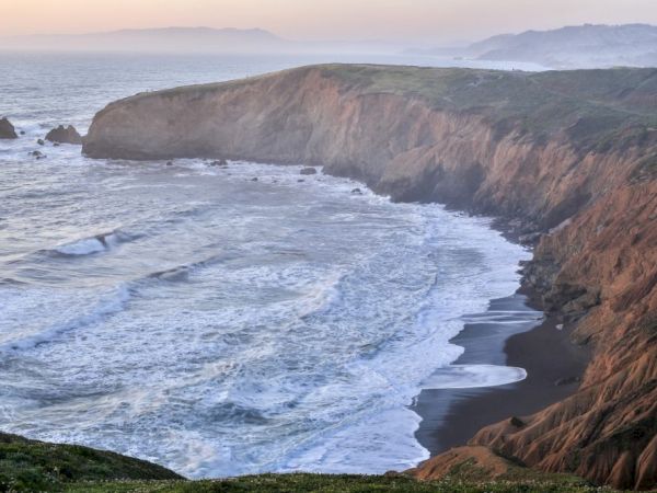 A scenic coastal landscape with cliffs, waves crashing on a sandy beach, and distant hills under a calm sky, creating a serene view.