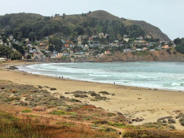 A coastal scene with a sandy beach, ocean waves, and a hillside with houses and trees. Overcast sky adds to the serene atmosphere.