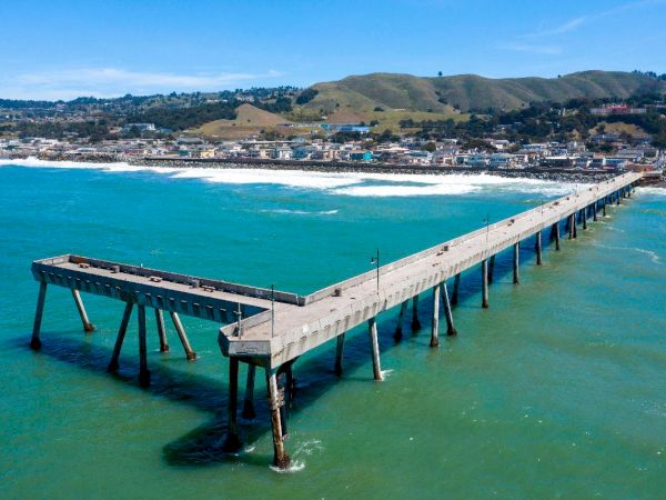 A long pier extends over turquoise water towards a coastal town with hills in the background, under a clear blue sky.