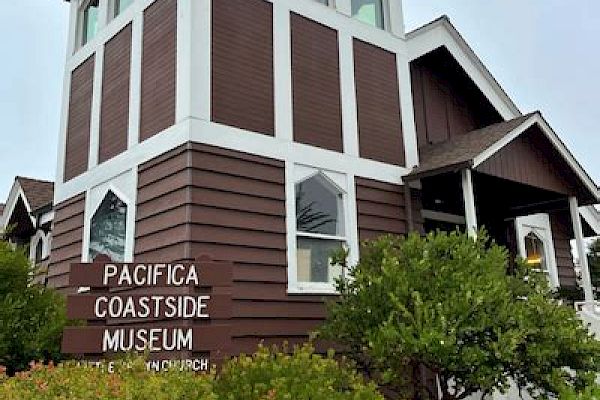 The image shows a brown and white building labeled "Pacifica Coastside Museum" surrounded by greenery and a cloudy sky.