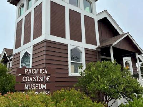 The image shows a brown and white building labeled "Pacifica Coastside Museum" surrounded by greenery and a cloudy sky.