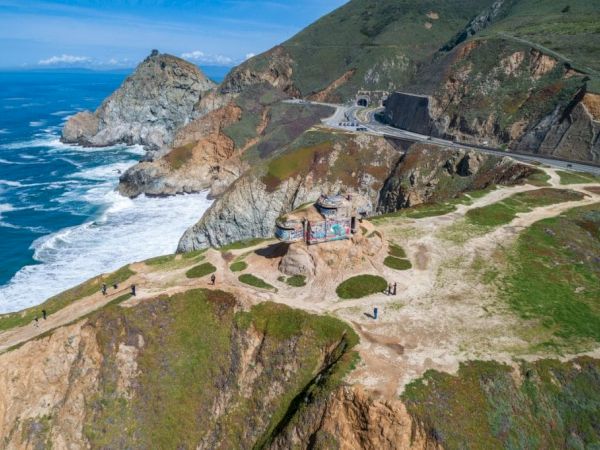 A coastal view shows a scenic overlook above cliffs and ocean waves, with a road leading through the rugged landscape under a blue sky.
