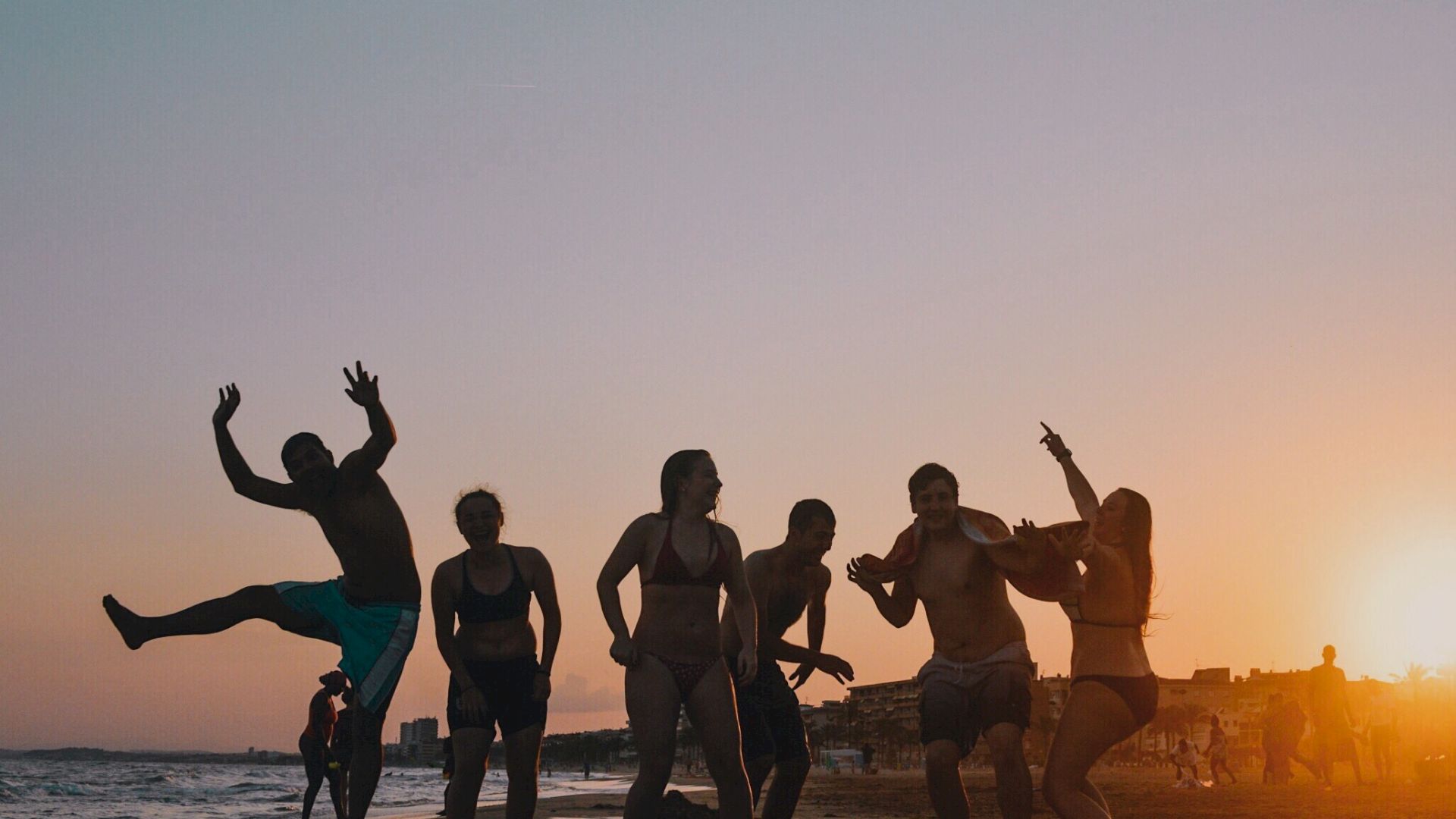 A group of people enjoying sunset on a beach, silhouetted against the colorful sky, with one person playfully jumping.