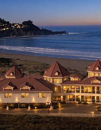 Warm-lit houses overlook a sandy beach and crashing waves at dusk, with hills and a glowing coastline in the background.