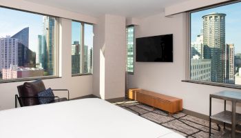 A modern hotel room with a bed, TV, chair, and cityscape view through large windows.