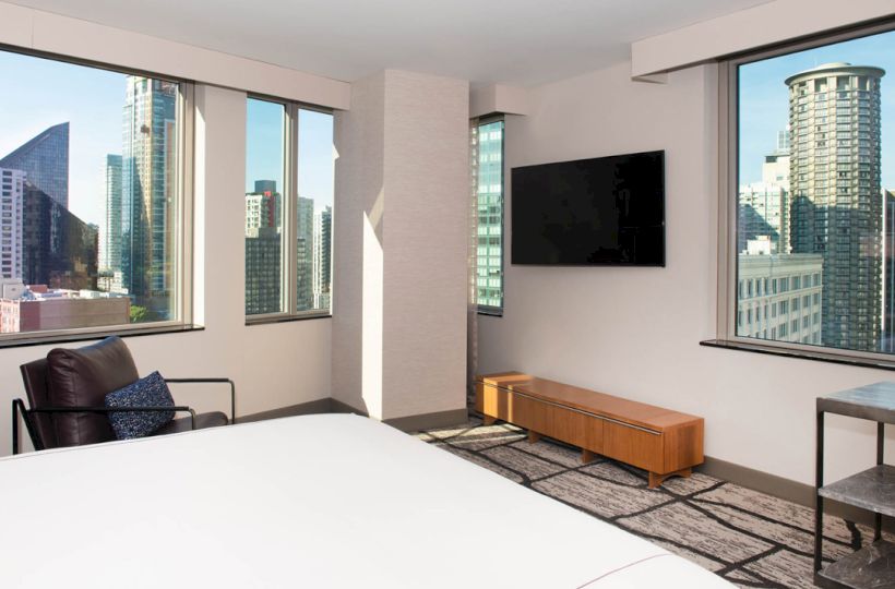 A modern hotel room with a bed, TV, chair, and cityscape view through large windows.