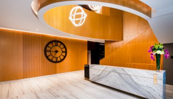 The image shows a modern lobby with wooden walls, a large clock, a marble reception desk, and decorative lights hanging from above.
