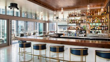 The image shows a stylish bar area with a wooden counter, modern bar stools, and shelves stocked with various bottles against a city view backdrop.
