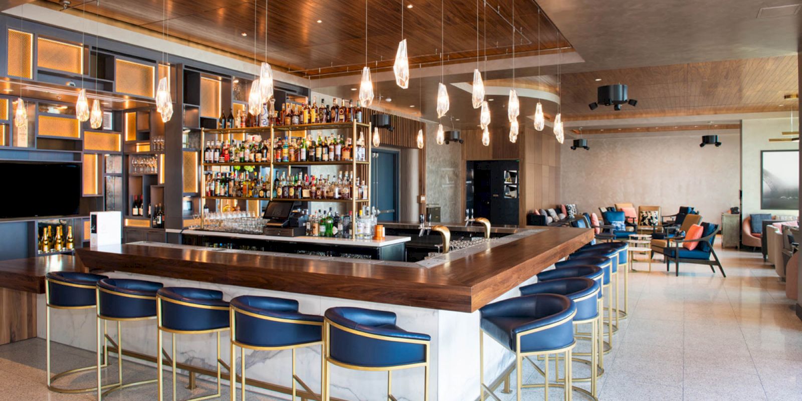 A modern bar with hanging lights, a stocked liquor shelf, blue stools, and a lounge area with chairs and tables in the background.