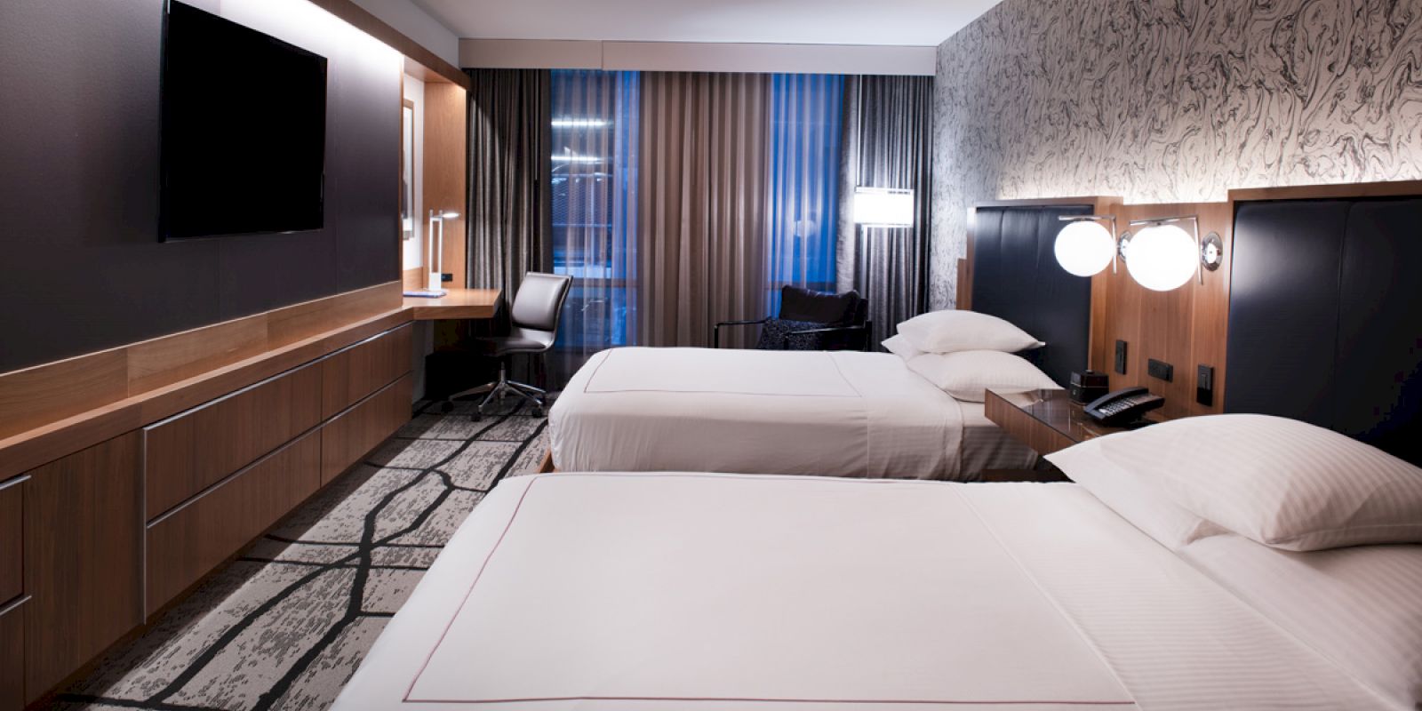 A modern hotel room with two beds, a TV, a desk, and a window with curtains. The decor features dark tones and stylish lighting.