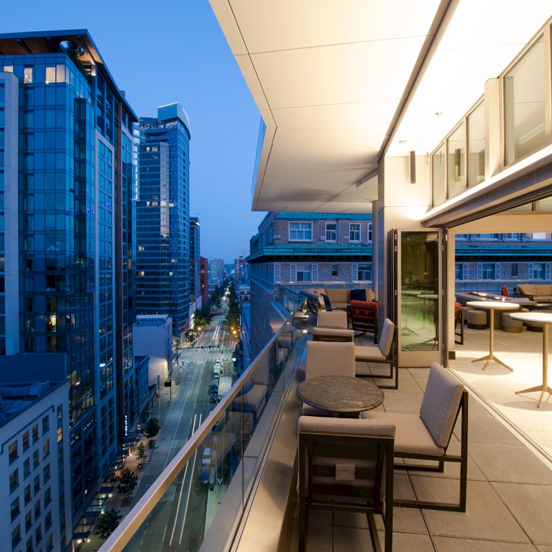 The image shows a modern cityscape with a high-rise building balcony, featuring chairs and tables, overlooking a street below at dusk.