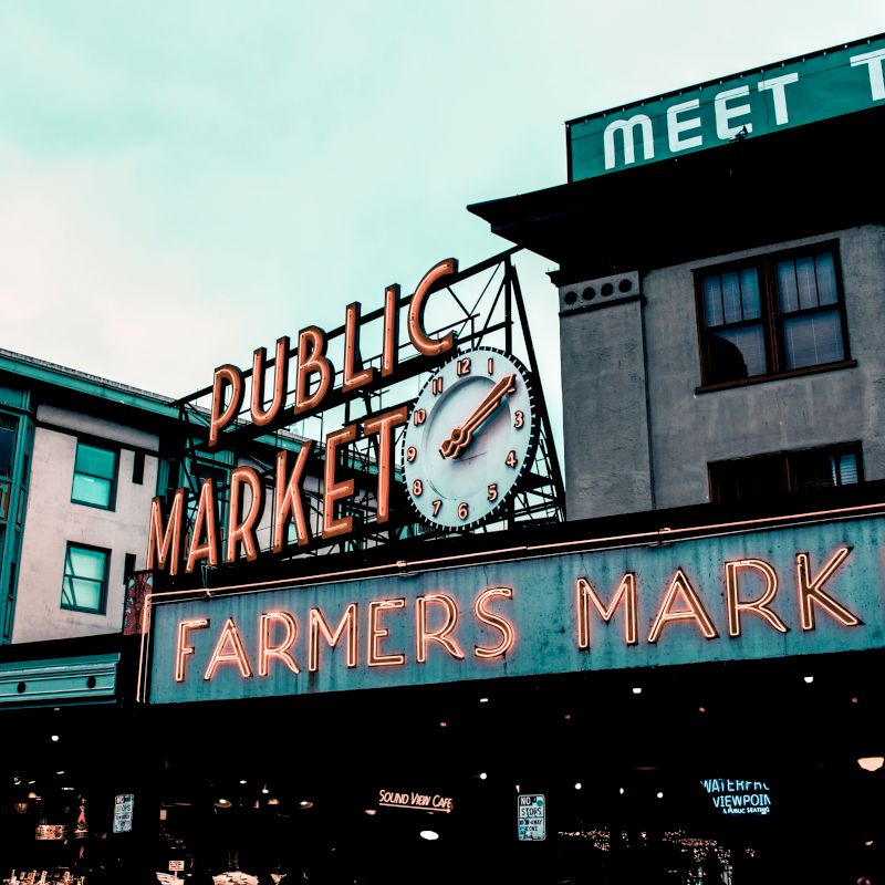 The image shows an iconic sign for a public market and farmers market, with neon lights and a clock, set against a cloudy sky.