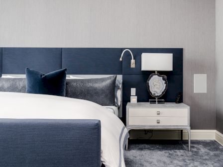 A modern bedroom with a navy headboard, white bedding, bedside table, lamp, and decorative items on a soft gray carpet.