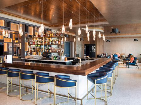 A modern bar with a wooden counter, blue stools, hanging lights, and a variety of bottles. Cozy seating in the background enhances the ambiance.