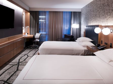 The image shows a modern hotel room with two beds, a wall-mounted TV, a desk with a chair, and patterned carpet and wallpaper.