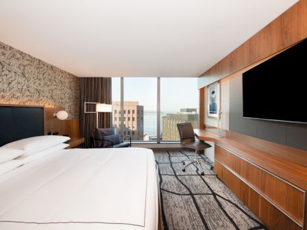 A modern hotel room with a large bed, flat-screen TV, desk, and city view through a wide window.